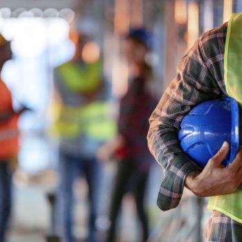 Man holding blue helmet close up. Construction man worker with office and people in background. Close up of a construction worker's hand holding working helmet.